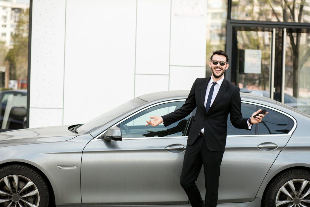 uber driver in elegant suit in front of an luxury car showing his cellphone, outside on the street in front of an office building