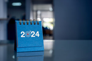 Happy new year 2024 background. 2024 year numbers with target goal icon on blue small desk calendar cover standing on glass table in office with copy space. Business goals and success concepts.