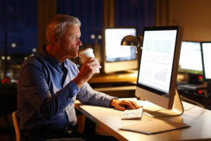 Portrait of senior financial advisor sitting at workplace in front of computer at late night and drinking coffee while analyzing data.