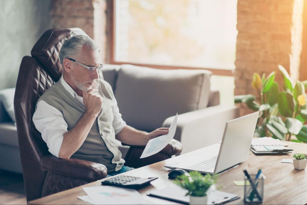 old man looking at his laptop while holding a paper